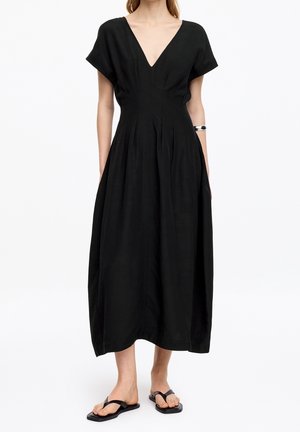 Mujer que lleva un vestido midi negro con mangas cortas y escote en V profundo combinado con sandalias planas negras y una pulsera plateada.