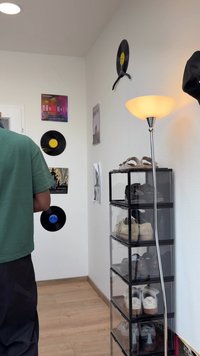 Fekete és áttetsző műanyag cipőtároló egység, egymásra rakott fiókokkal, különböző lábbelikkel. A falon vinyl lemezek és művészeti alkotások láthatók. A padló lámpa meleg fényt áraszt.