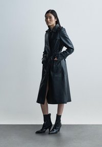Czarny płaszcz skórzany z kołnierzem, przednimi kieszeniami i paskiem w talii. Modelka nosi czarne botki na szpilce. Gładka faktura.