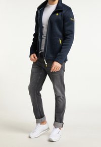 Sweat à capuche zippé bleu marine avec un tissu texturé, des fermetures éclair accent jaunes et un logo brodé. Associé à un jean gris retroussé et des baskets blanches.