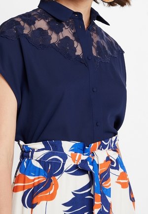 Marineblaue Bluse mit Spitzenblumenpasse und Knöpfen, kombiniert mit einem hoch taillierten Rock mit abstraktem Blumenmuster in Blau, Orange und Weiß und einem gebundenen Gürtel.