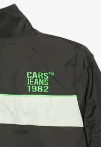 Chaqueta en blanco y negro por detrás con el texto en verde "CARS JEANS 1982" y una franja blanca horizontal en la parte superior de la espalda.