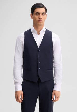 Uomo che indossa un gilet blu navy con bottoni sopra una camicia bianca e pantaloni blu navy abbinati, in piedi davanti a uno sfondo semplice.