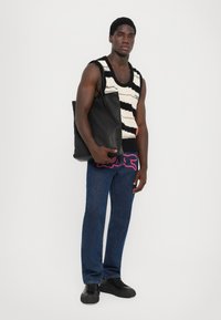 Sac fourre-tout noir au design épuré, fabriqué en matériau lisse. Le modèle porte un débardeur en tricot à rayures avec des bretelles larges et un jean en denim foncé.