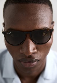 Ochelari de soare Ray-Ban cu rame în stil țestoasă și lentile închise la culoare, cu o formă distinctivă. Prim-plan al feței unei persoane, evidențiind pielea netedă și un fundal neutru.