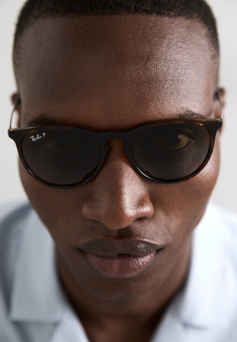 Ochelari de soare Ray-Ban cu rame în stil țestoasă și lentile închise la culoare, cu o formă distinctivă. Prim-plan al feței unei persoane, evidențiind pielea netedă și un fundal neutru.