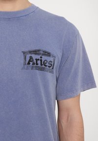 Sinine puuvillane T-särk, mille vasakul rinnal on must trükitud logo "Aries" stiilse arhitektuuri disainiga. Lühikesed varrukad.