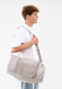 Grau gefärbte Sporttasche aus strukturiertem Stoff, mit zwei Griffen, einem langen Schulterriemen und einem Etikett auf der Vorderseite.