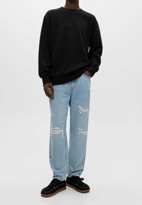 Persona che indossa un felpa nera a maniche lunghe, jeans strappati azzurri chiari e scarpe nere con lacci su uno sfondo bianco uniforme.
