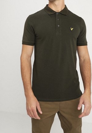 Man wearing a dark green short-sleeve polo shirt with a yellow bird logo on the chest and brown pants, standing against a plain background.