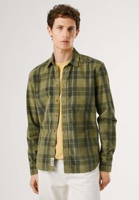 Grønn rutete flanellskjorte med mørk grønne og beige ruter, knapping foran, og en patch-label i kanten. Bært over en gul skjorte.