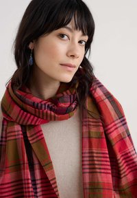 Rødt og grønt ternet tørklæde med varierende stribemønstre, hængende løst omkring halsen. Blød tekstur, levende farver og lagdelt design.