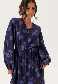 Flödande klänning med V-ringning i mörkblått med blommönster i ljusblått, utrustad med långa ärmar och knuten midja för en definierad siluett.