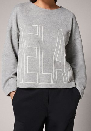 Person trägt ein graues Sweatshirt mit großen, umrandeten Buchstaben, die "RELAX" buchstabieren, kombiniert mit schwarzen Hosen und Händen in den Taschen.