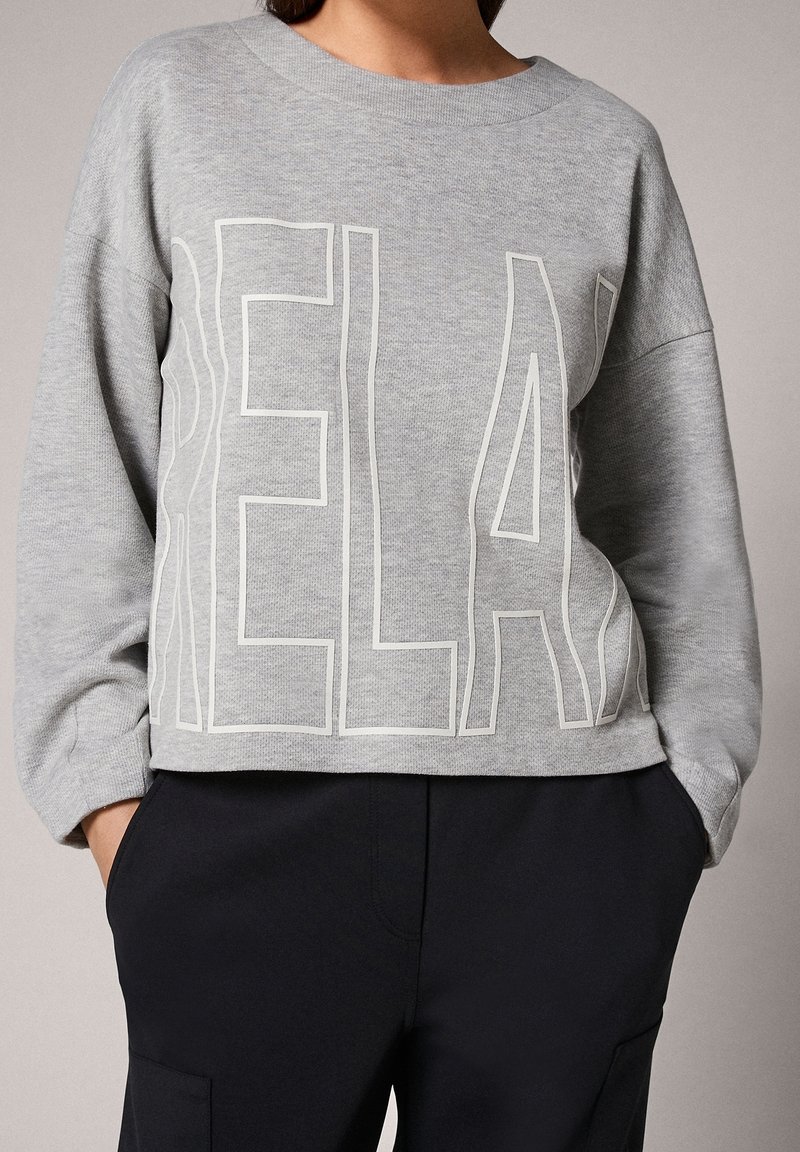 Personne portant un sweat-shirt gris avec de grandes lettres en contour formant le mot « RELAX », associée à un pantalon noir et les mains dans les poches.