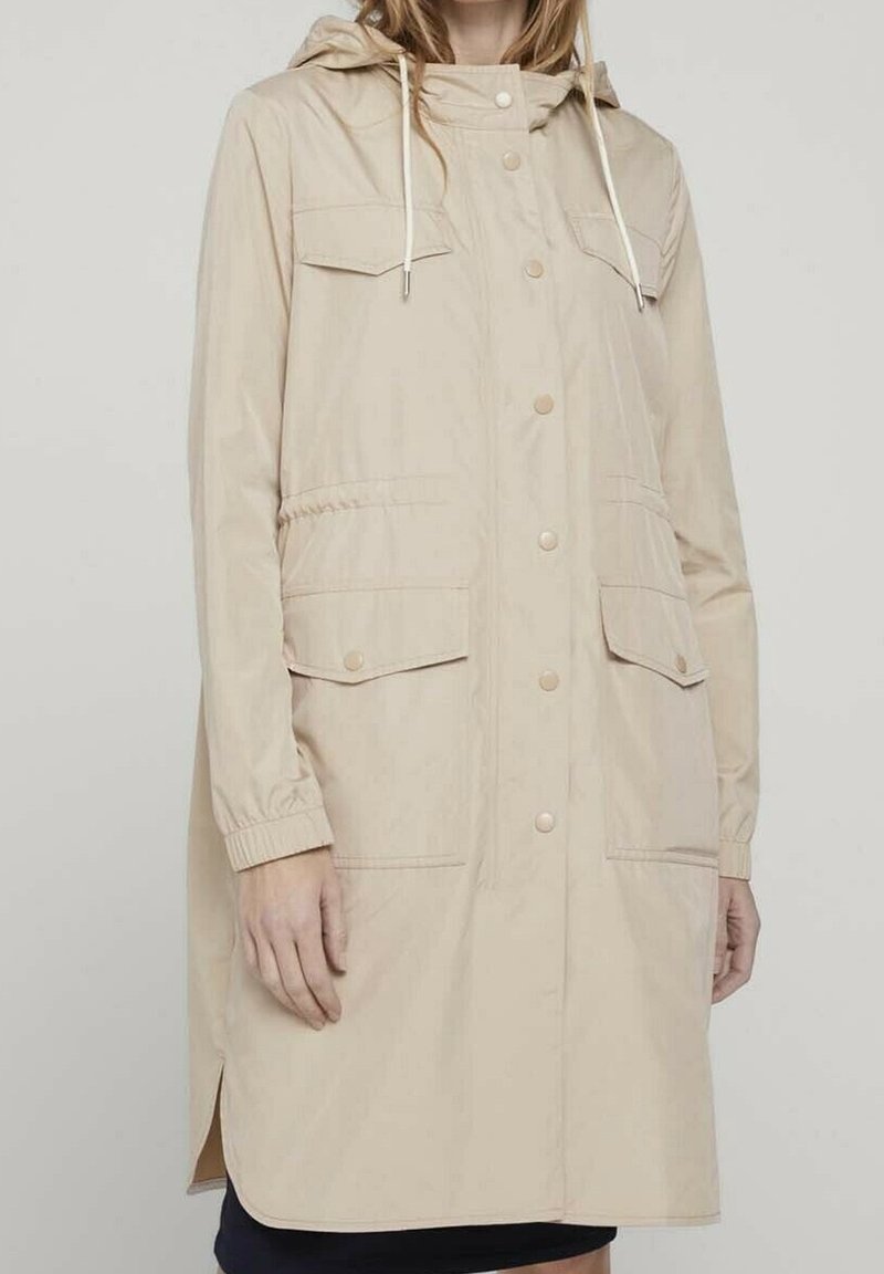 Abrigo impermeable beige con capucha, cierre frontal de botones a presión, dos bolsillos en el pecho, puños elásticos y aberturas laterales para mayor facilidad de movimiento.