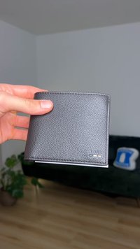 Sort læderpung med en tekstureret overflade, med et lille præget logo og et dobbelt foldedesign, holdt i en hånd mod en sløret baggrund.