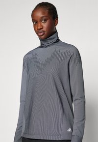 Pilkas golfas su tekstūruotu dizainu, turinčiu vertikalias linijas ir ilgomis rankovėmis. Apatinėje dalyje yra Adidas logotipas.
