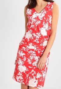 Czerwona sukienka w kwiaty z dekoltem w kształcie litery V, bez rękawów, z marszczeniem w talii. Posiada białe abstrakcyjne wzory kwiatowe na gładkim materiale.