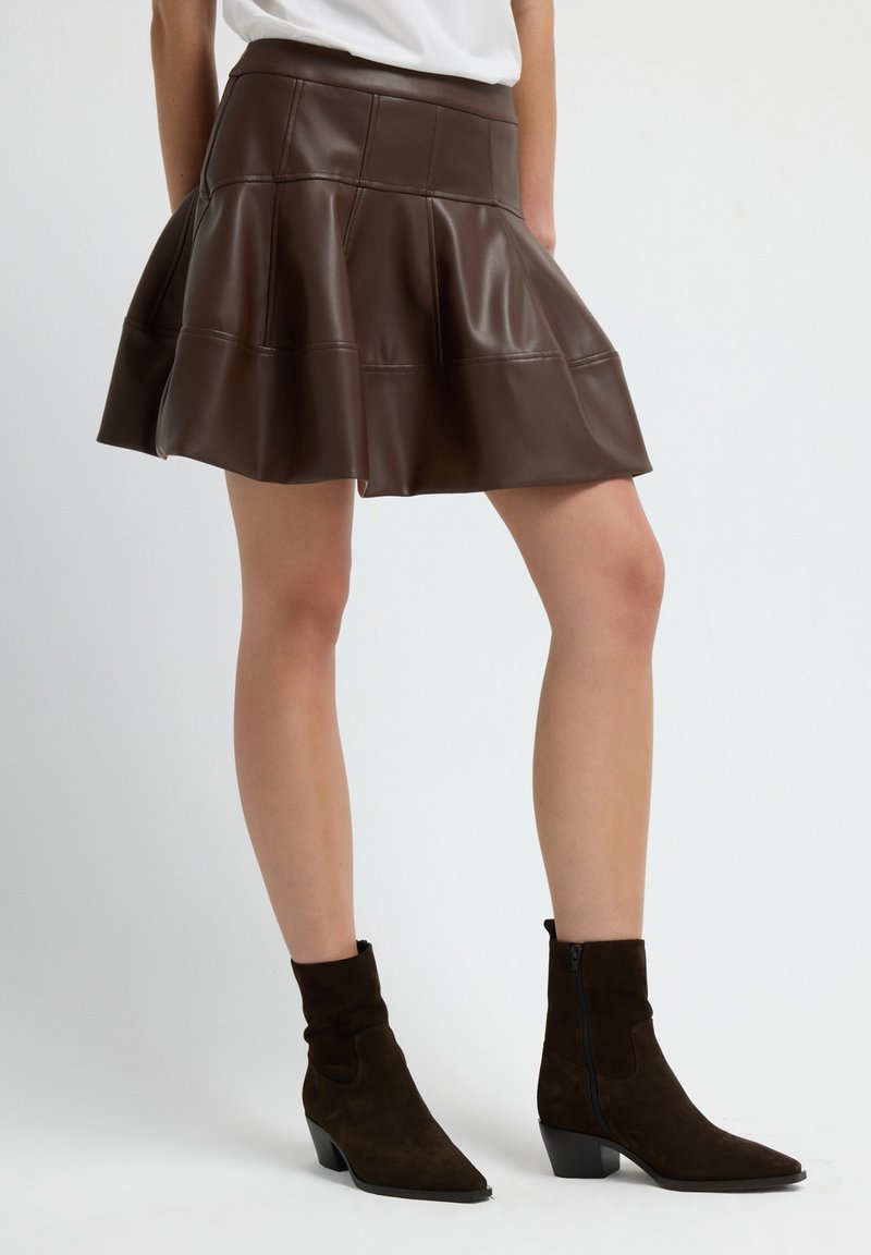 Jupe évasée en simili cuir marron avec une taille ajustée, présentant des détails de couture. Associée à des bottines en daim marron à bout pointu.