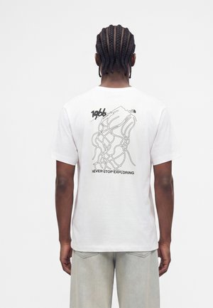 Άνδρας με πλεξούδες που φοράει λευκό T-shirt με μαύρο αφηρημένο σχεδιασμό γραμμής, "1966," και "Never Stop Exploring" κείμενο στην πλάτη.