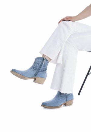 Botines de ante azul con un diseño decorativo perforado, tachuelas plateadas y un tacón bajo de madera, combinados con pantalones blancos acampanados.