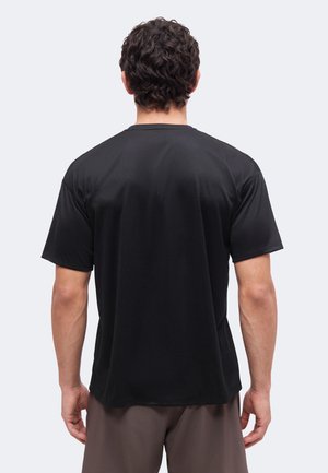 Homme aux cheveux bouclés et foncés portant un t-shirt noir uni à manches courtes et un pantalon marron, debout, dos à un fond clair.