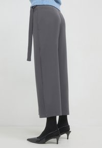 Femme portant un pantalon cropped large gris avec une ceinture en tissu, des collants noirs et des escarpins à talon aiguille noirs sur fond blanc.