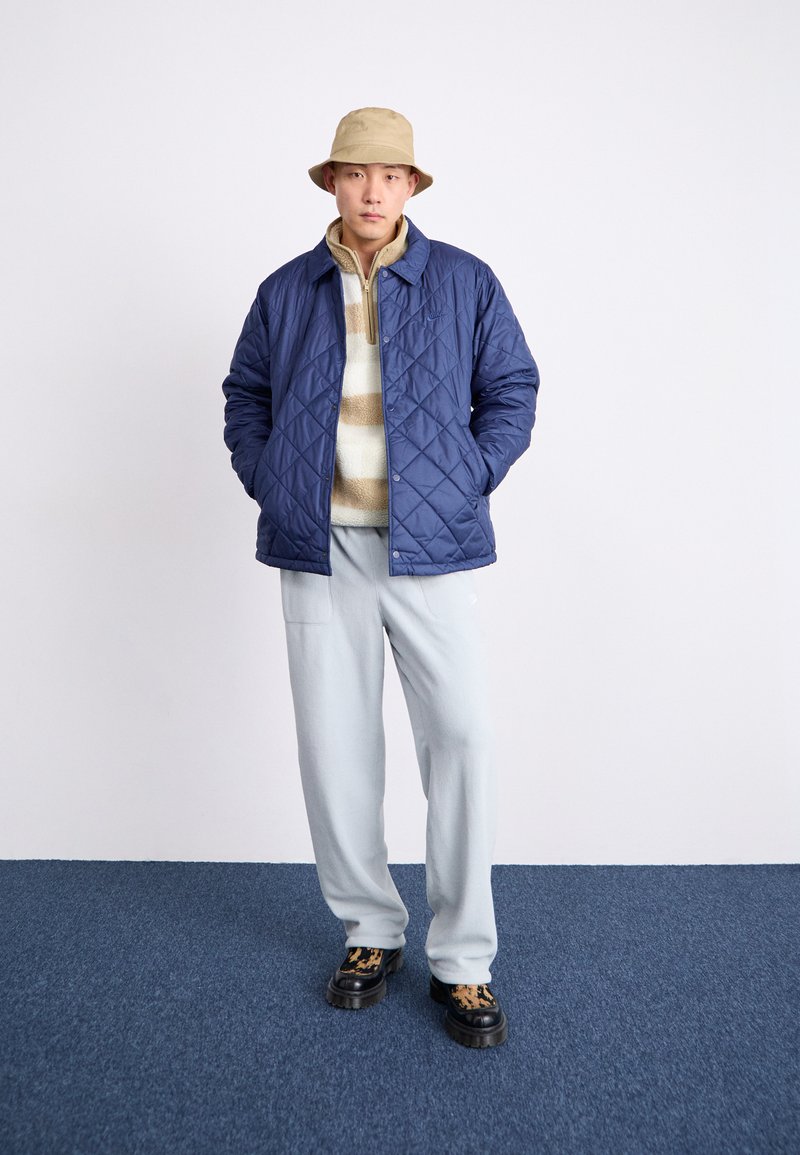 Veste matelassée bleu marine sur un pull en polaire rayé beige et blanc, assortie avec un pantalon bleu clair et des chaussures noires à motif camouflage marron.