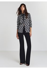 Schwarz-weiße gemusterte Bluse mit geometrischen Designs, langen Ärmeln und Kragen, kombiniert mit schwarzen Glockenhosen und Riemchen-Absätzen.