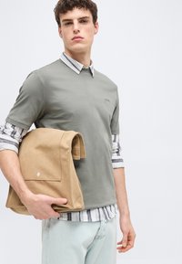 Νεαρός άνδρας που φοράει ριγέ πουκάμισο με στρώσεις και γκρι μπλουζάκι, κρατώντας διπλωμένο μπεζ σακάκι μπροστά σε απλό φωτεινό φόντο.