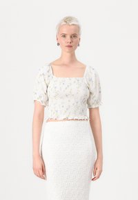 Bijeli crop top s cvjetnim uzorkom, kvadratnim izrezom i puf rukavima. Teksturirana tkanina s volanima na rubu. Uparen s bijelom suknjom.