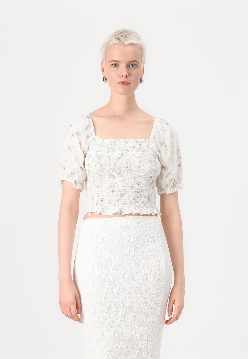 Bijeli crop top s cvjetnim uzorkom, kvadratnim izrezom i puf rukavima. Teksturirana tkanina s volanima na rubu. Uparen s bijelom suknjom.