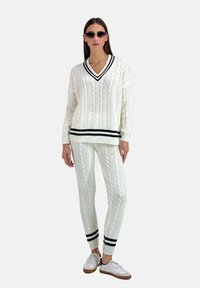 Gebreide trui en legging set in gebroken wit met zwarte strepen, met kabelgebreide structuur en v-hals ontwerp, gecombineerd met witte sneakers.