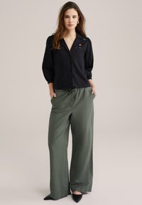 Camicia nera con colletto e maniche a palloncino, abbinata a pantaloni a gamba larga verde oliva. Scarpe nere con punta a stiletto.