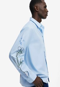 Light blue button-up shirt with long sleeves featuring floral embroidered design on the sleeve and subtle texture.