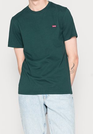 Mężczyzna ubrany w ciemnozielony T-shirt Levi's i jasnoniebieskie dżinsy, z widocznym tatuażem na lewym przedramieniu.