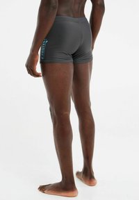 Graue Badeshorts mit engem Sitz, versehen mit einem blauen "Protest"-Logo an der Seite, aus glattem, leichtem Material gefertigt.