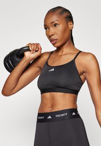 Svart sportsbh med tynne stropper, som har en glatt tekstur, et logo foran og en støttende kant; kombinert med svarte høyhånds leggings.