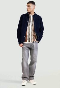 Jovem vestido com uma jaqueta preta abotoada sobre uma camisa estampada, jeans cinzentos e ténis brancos, de pé com uma mão no bolso.