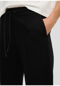 Zwarte broek met een gestructureerde afwerking, voorzien van een trekkoord in de taille, zijzakken en een getailleerde pasvorm. Close-up weergave die de ontwerpdDetails benadrukt.