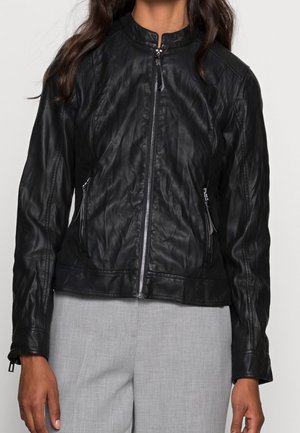 Veste en cuir noire avec fermeture éclair devant, col montant et poches latérales zippées portée sur un pantalon gris clair.
