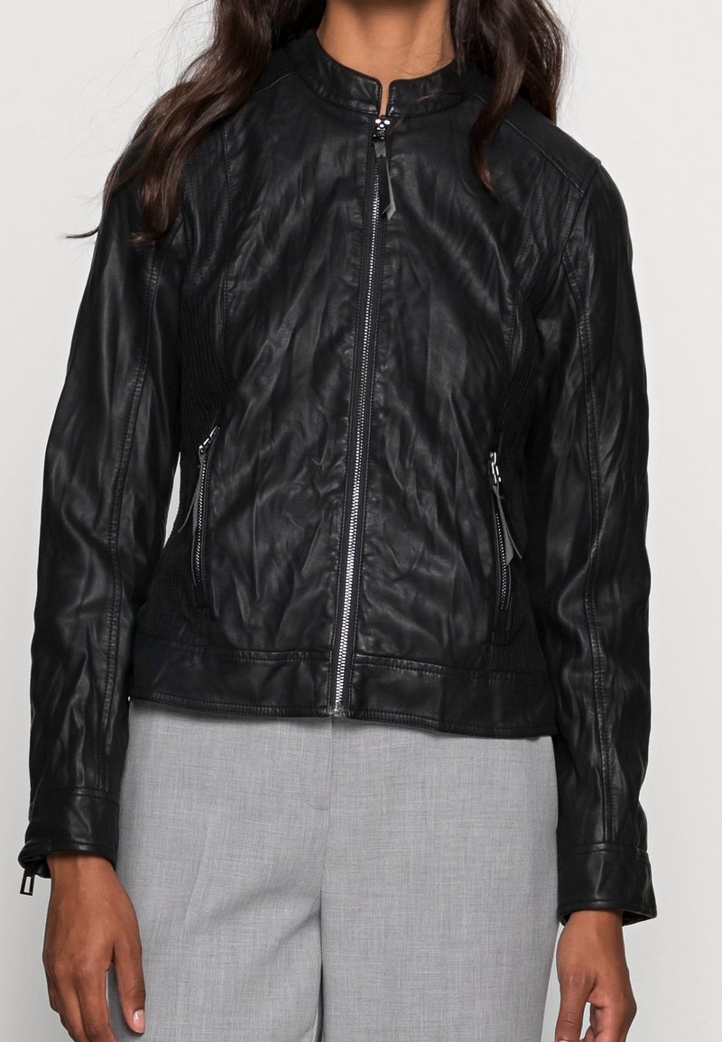 Veste en cuir noire avec fermeture éclair devant, col montant et poches latérales zippées portée sur un pantalon gris clair.