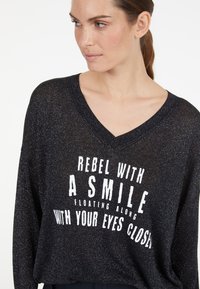 Schwarzer, lässig geschnittener Pullover mit V-Ausschnitt, der eine glänzende Textur aufweist. Frontdruck in Weiß mit dem Text: "REBEL WITH A SMILE FLOATING ALONG WITH YOUR EYES CLOSED."