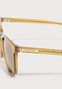 Óculos de sol com lentes de tonalidade castanha e armação âmbar transparente. Apresenta dobradiça metálica nos braços e logótipo "Calvin Klein" na haste. Forma quadrada.