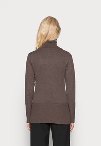 Femme blonde vue de dos portant un pull à col roulé marron ajusté et un pantalon noir sur un fond clair uni.
