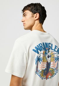 Camiseta de algodón blanca con un gráfico colorido de sirena en la parte posterior, rodeado de palmeras y el texto "WRANGLER" en azul.