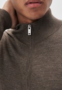 Rudas zip-up megztinis su aukštu apykakle, pagamintas iš tekstūruoto mezginio audinio. Yra sidabrinė užtrauktuko auselė su išgraviruotu „M“. 