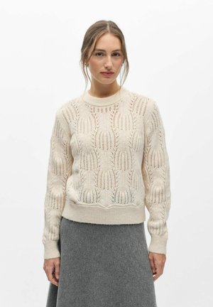 Femme portant un pull en maille crème à motif et une jupe en laine grise, debout devant un fond blanc uni.