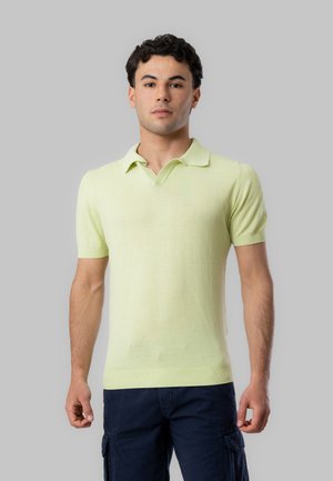 Giovane uomo in piedi rivolto in avanti, indossa una polo verde chiaro a maniche corte e pantaloni cargo blu scuro su sfondo grigio.